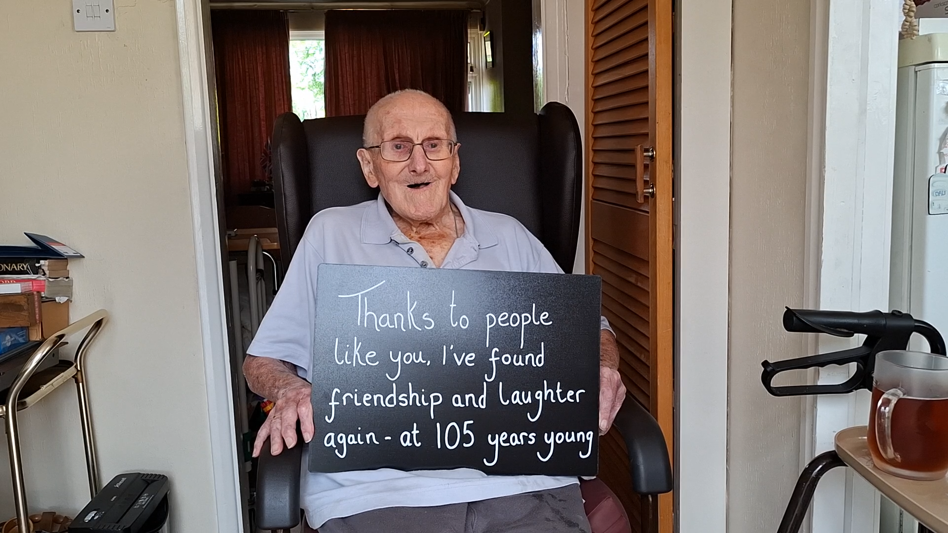 George in his home, sitting in a chair and holding a sign that reads 'Thanks to people like you, I've found friendship and laughter again - at 105 years young'