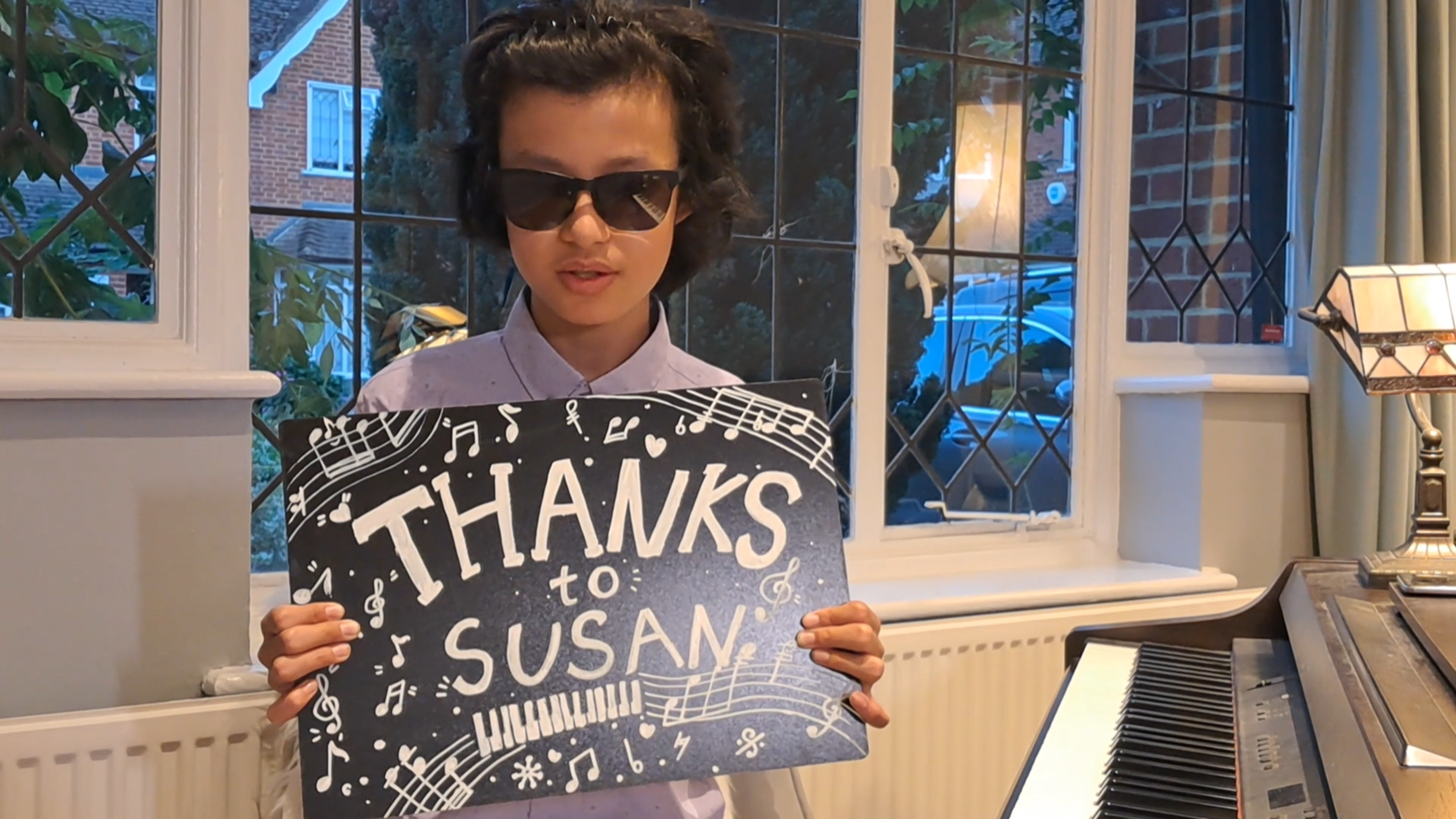 Chapman, sitting at his piano in his home, holding a sign that reads 'Thanks to Susan', surrounded by drawn musical notes