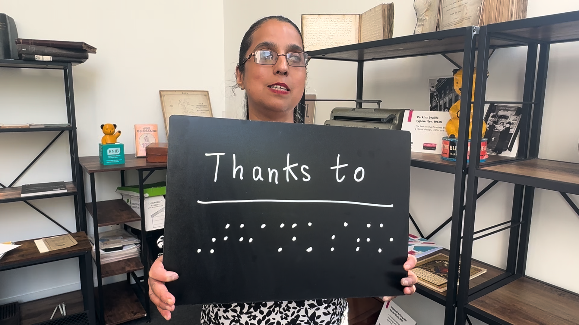 Amrit standing in an office with shelves around her, holding a sign that reads 'Thanks to...' followed by braille writing.
