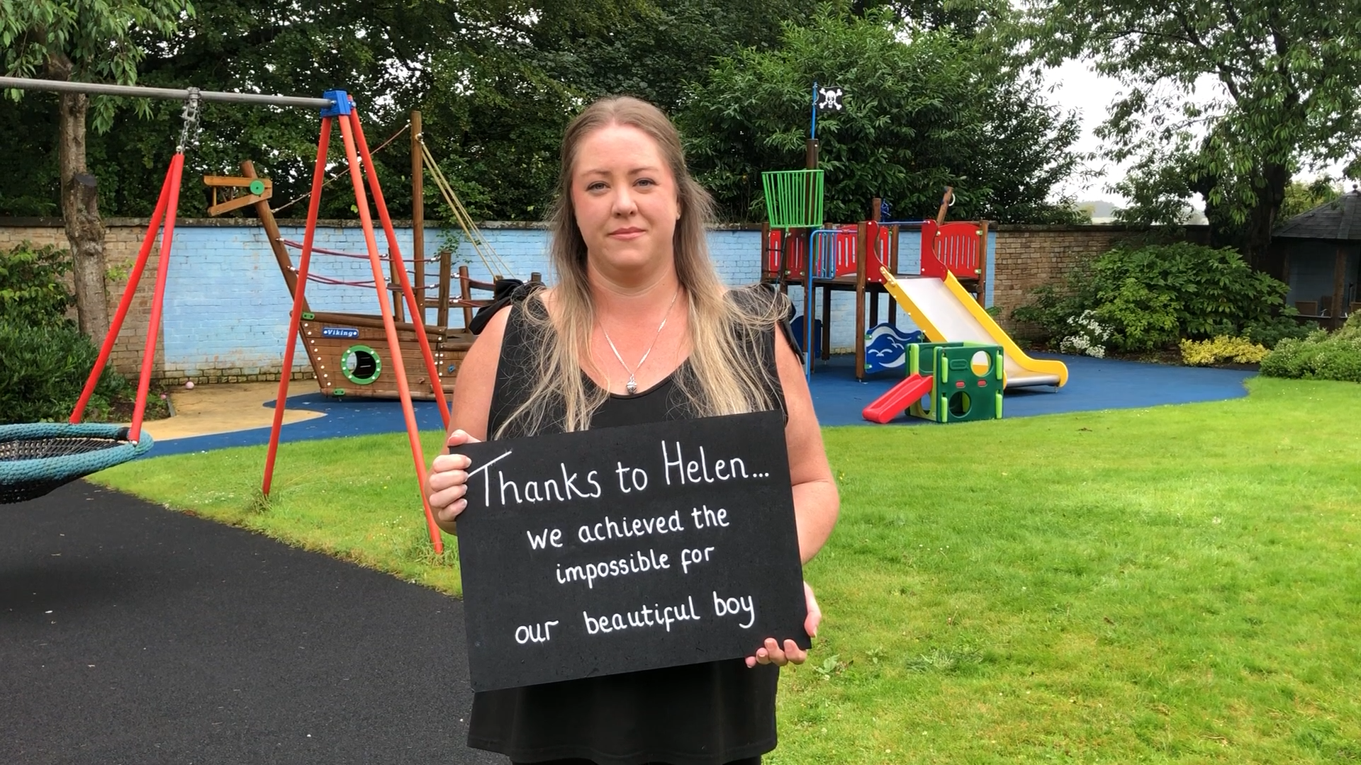 Cassy in the playground of Rachel House, holding a sign that reads 'Thanks to Helen, we achieved the impossible for our beautiful boy'